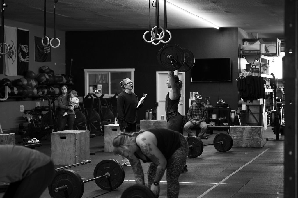 A woman lifting weights overhead in a gym setting, with other people and equipment visible.