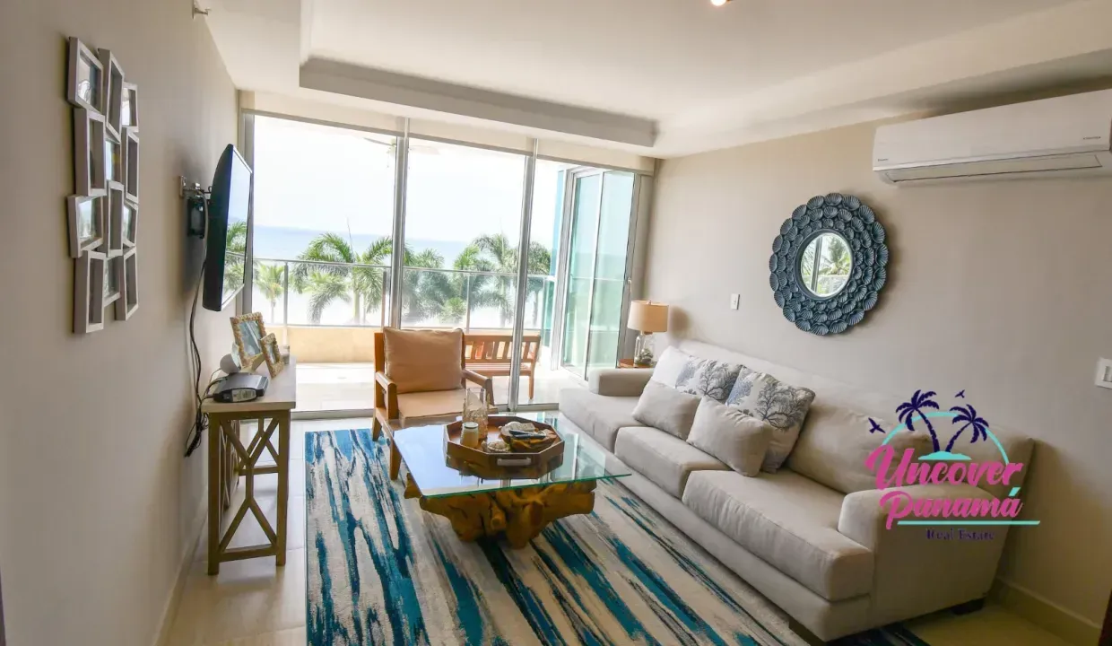 Bright living room with white sofa, blue striped rug, ocean-view windows, and a small dining table.