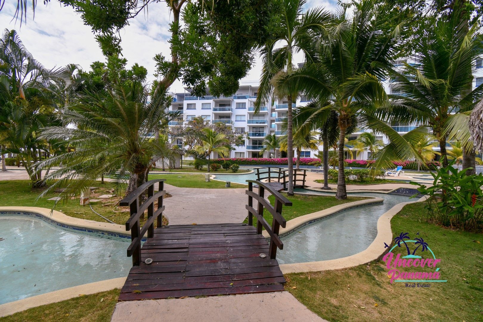 Wooden footbridge over a garden pond with palm trees and a white resort building in the background