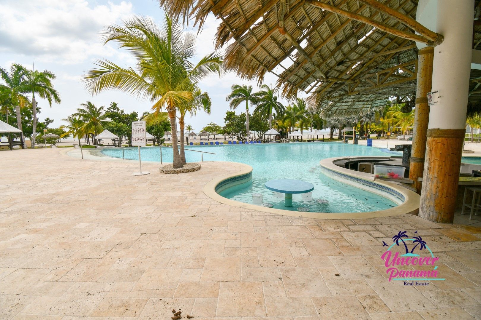 Tropical resort pool with palm trees, lounge chairs, and thatched-roof shade by the ocean