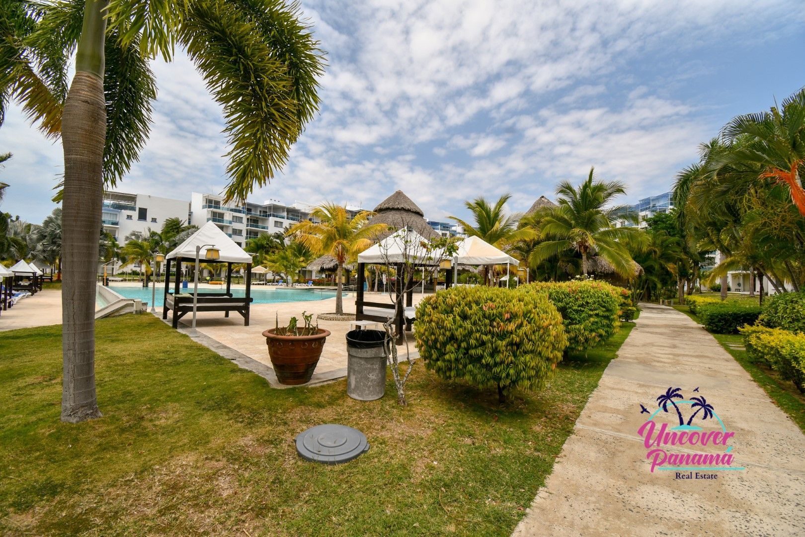Tropical resort garden with gazebos, palm trees, and a paved walkway under a cloudy sky.