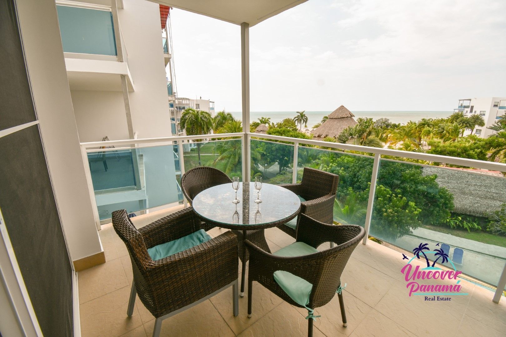 Balcony with wicker chairs and glass table overlooking lush greenery and the sea