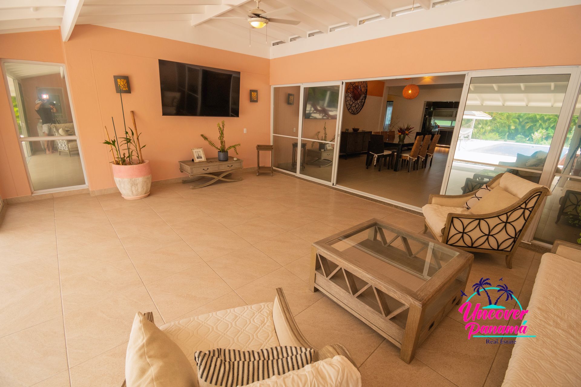 Covered patio seating area with beige furniture, wall-mounted TV, plants, and sliding glass doors to a dining room.