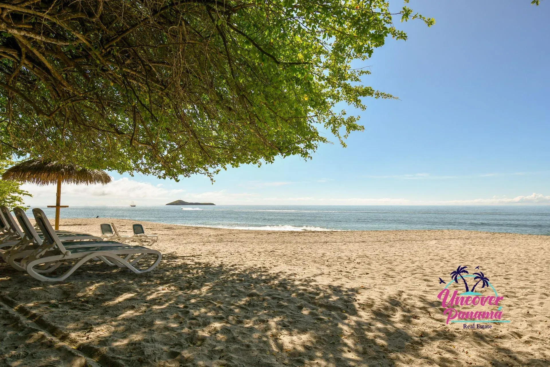 Shaded beach with lounge chairs, calm blue sea, and a distant island under a clear sky