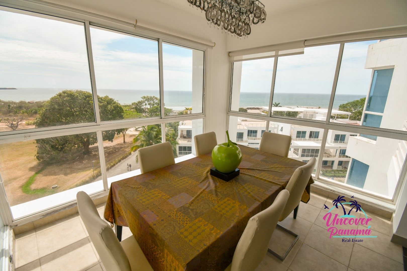 Bright dining area with a wooden table, beige chairs, and large windows overlooking the ocean and nearby buildings