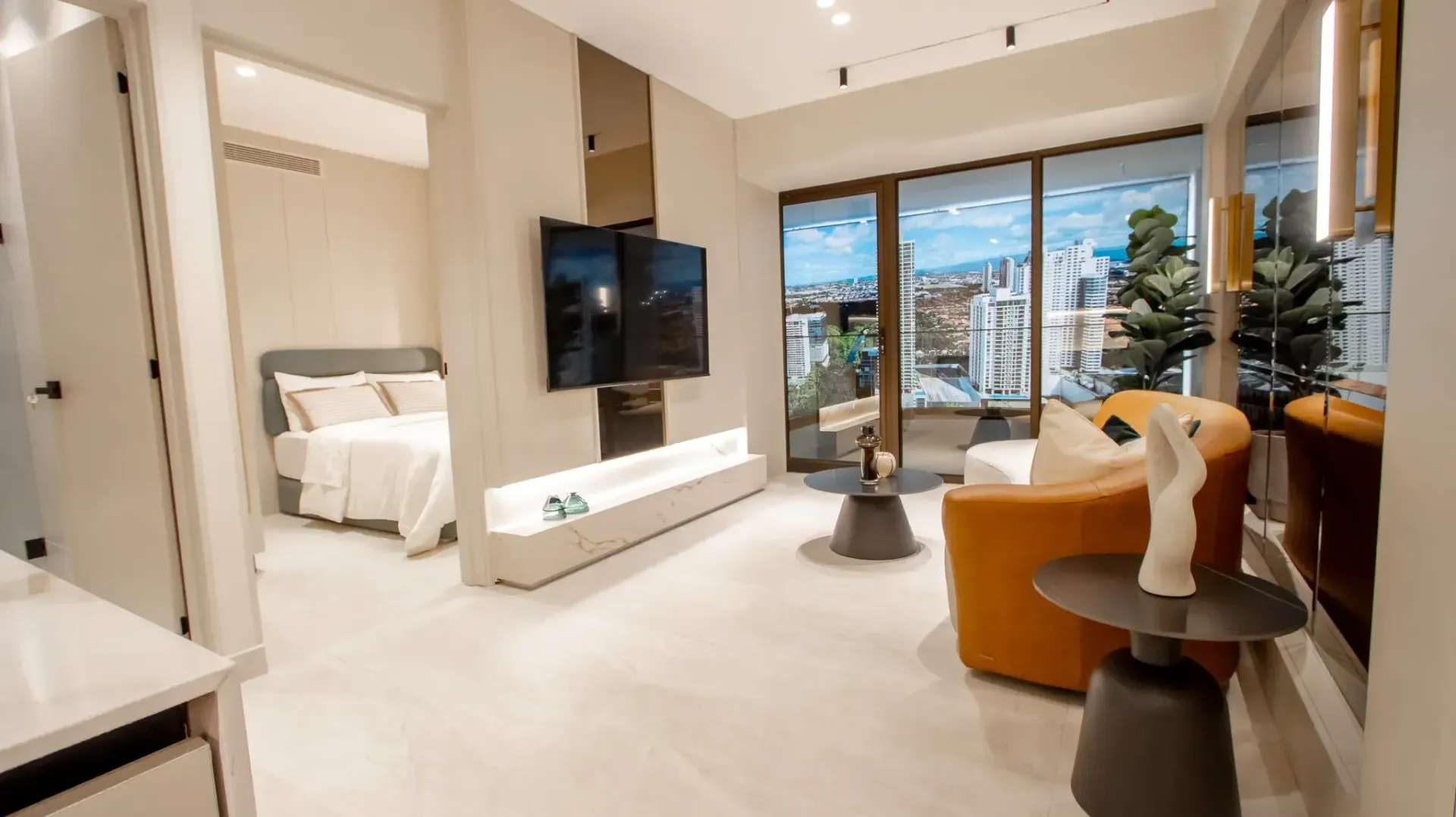 Modern apartment living room with large windows, sofa, TV, and bedroom visible through an open doorway