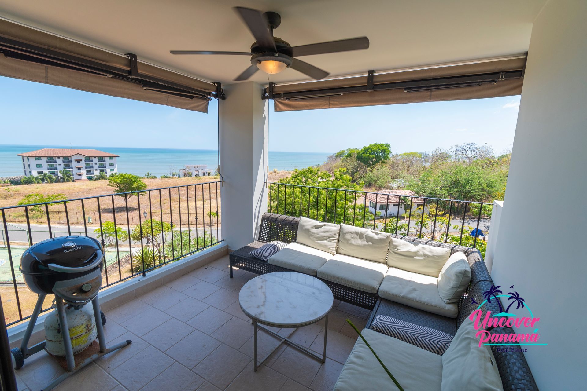 Covered patio with outdoor sofa, grill, and round table overlooking a grassy landscape and blue water.