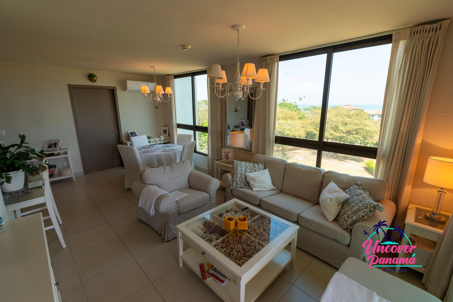 Bright living room with white sofas, coffee table, chandelier, and large windows overlooking a sunny view
