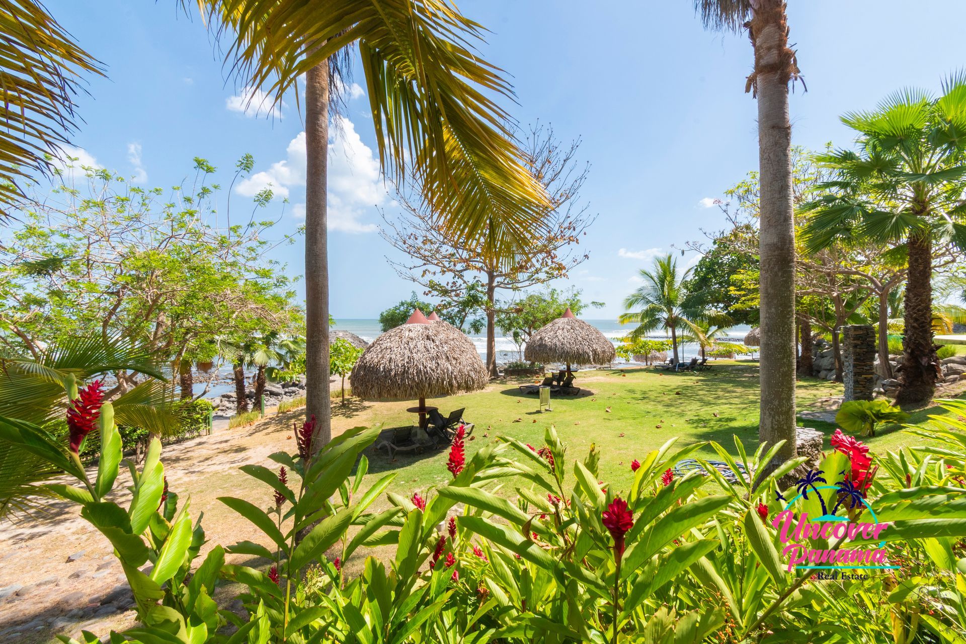 Tropical beach garden with palm trees, green plants, thatched huts, and blue sky by the shore