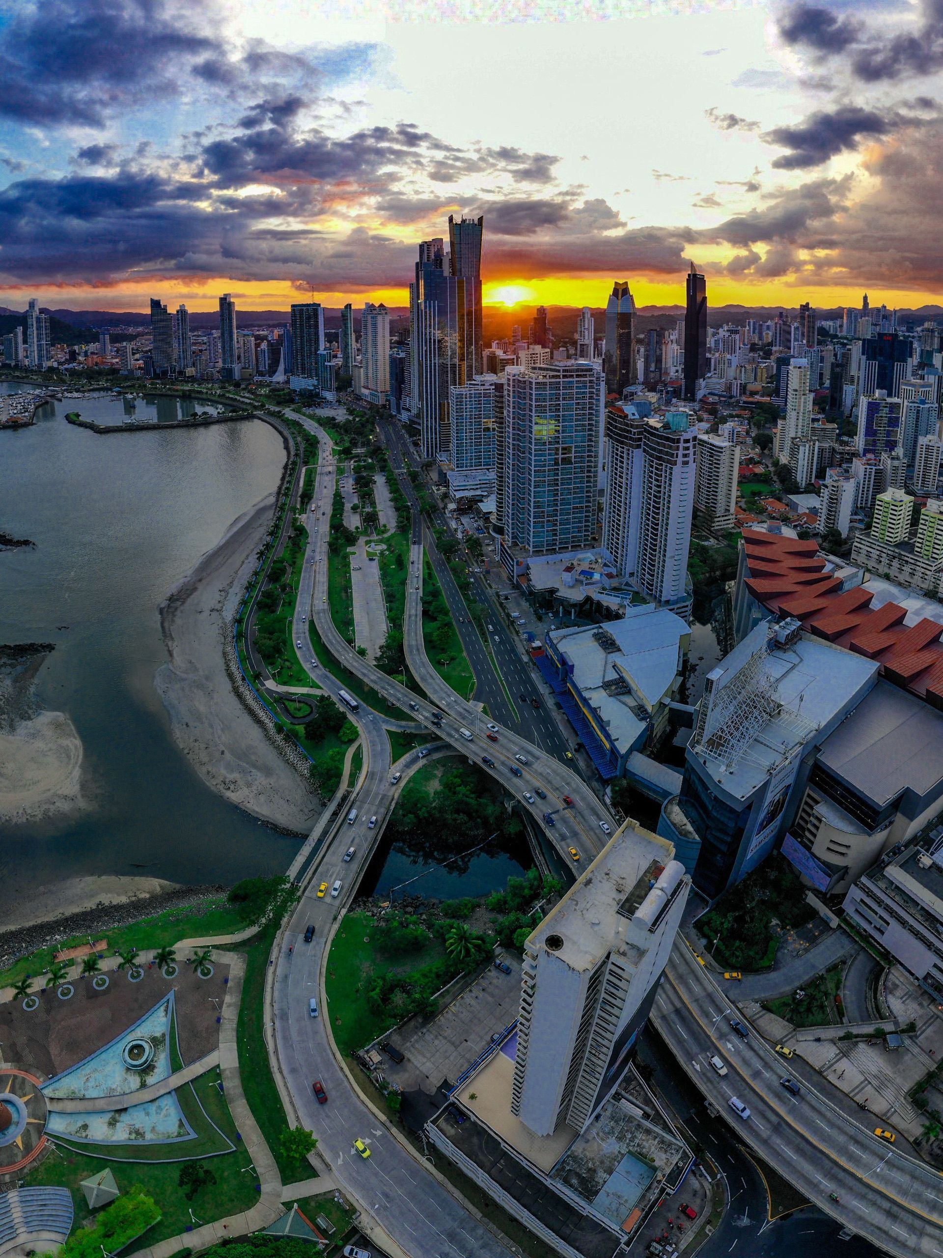 Aerial sunset view of a coastal city skyline, waterfront park, and curving roads beside the bay.