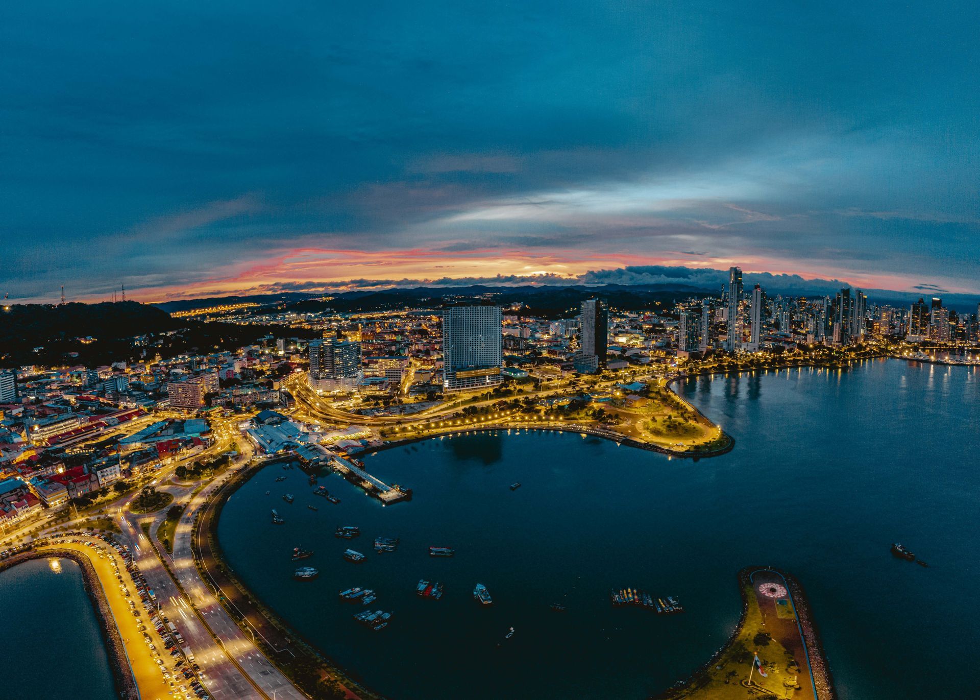 Dusk cityscape with a lit waterfront, harbor, and skyline under a blue-pink sky.