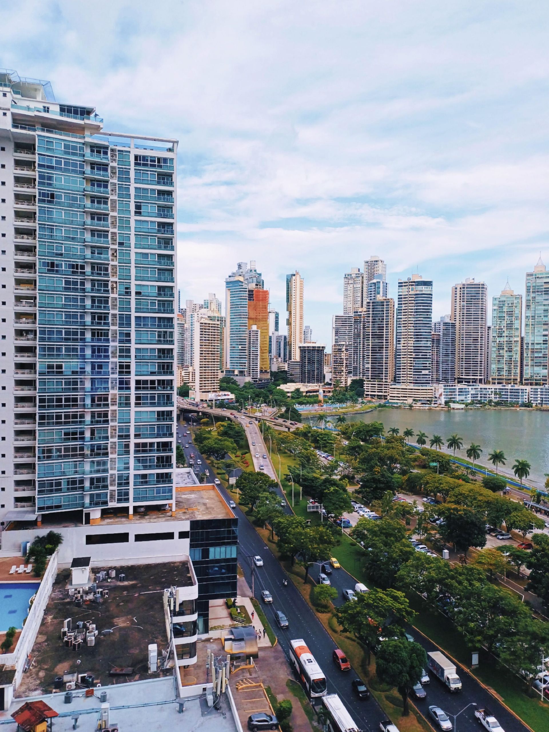 High-rise waterfront city skyline beside a tree-lined road and bay under a cloudy sky