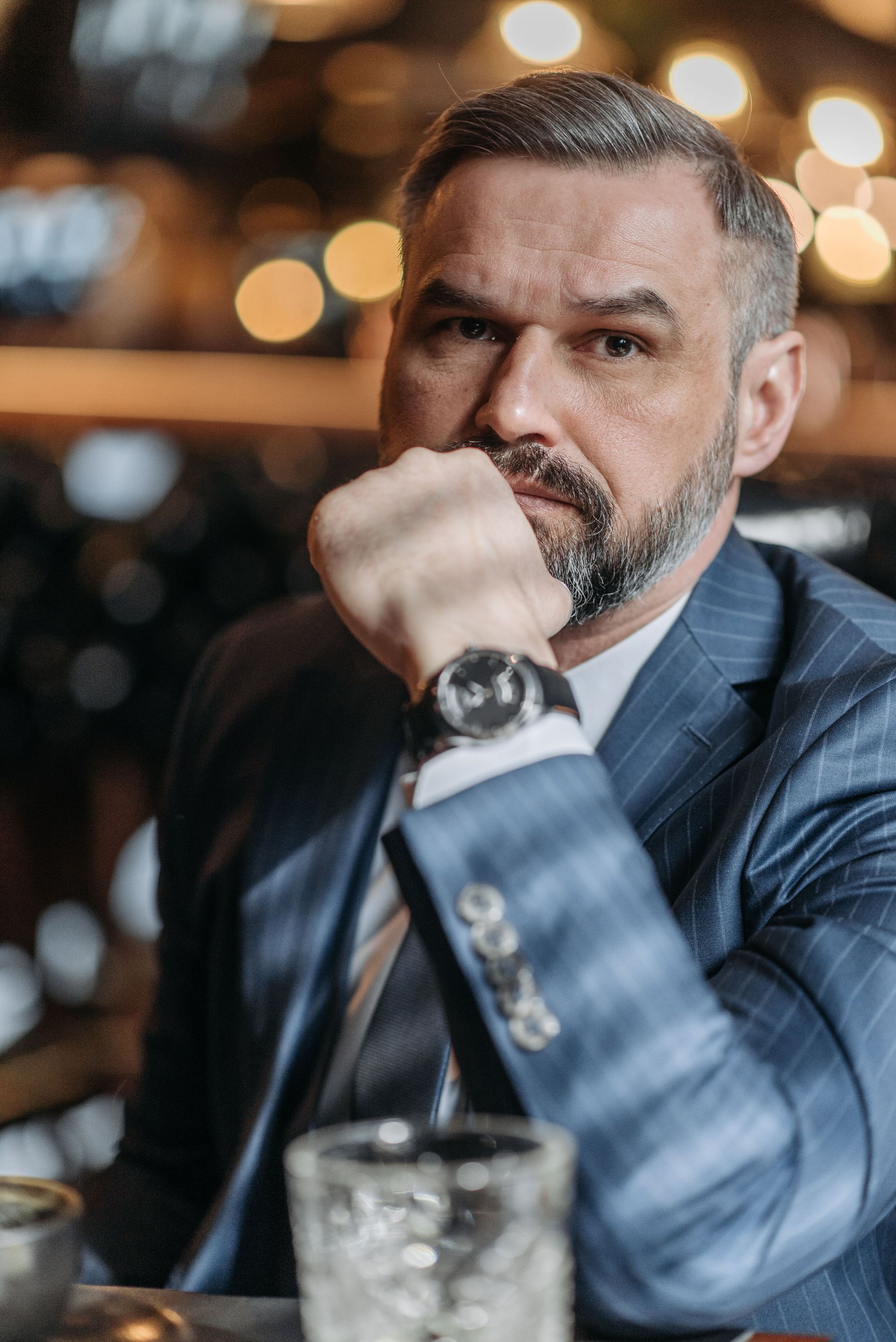 Man in blue suit resting his chin on his fist at a bar, with blurred warm lights behind him