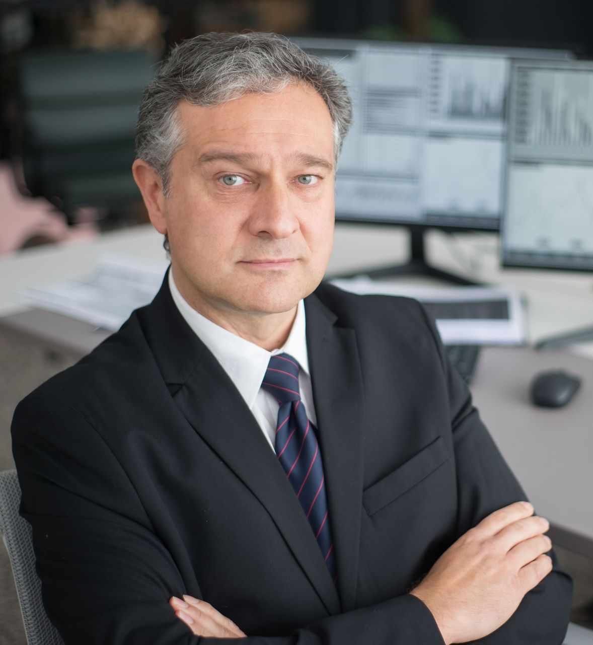 Businessman in a black suit with arms crossed in an office, looking at the camera.