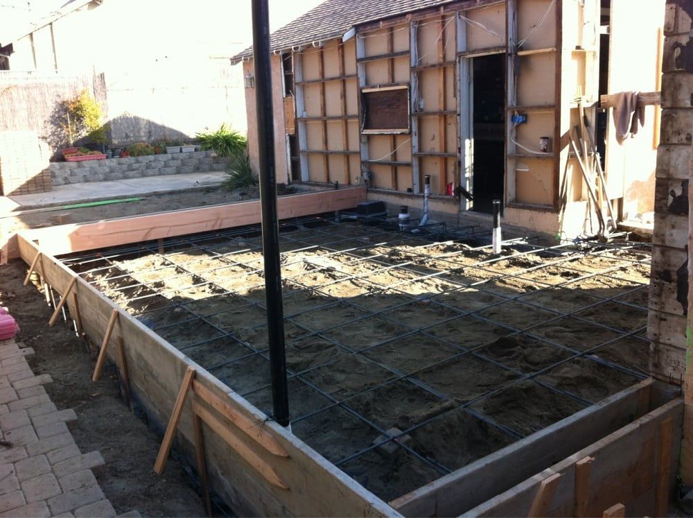 Concrete foundation under construction beside a house, with rebar grid and wooden framing.