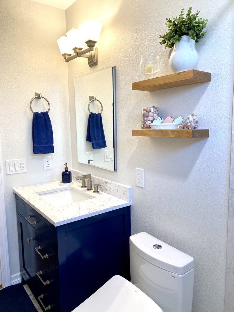 Small bathroom with dark vanity, white sink, toilet, mirror, towel rings, and floating shelves with decor.