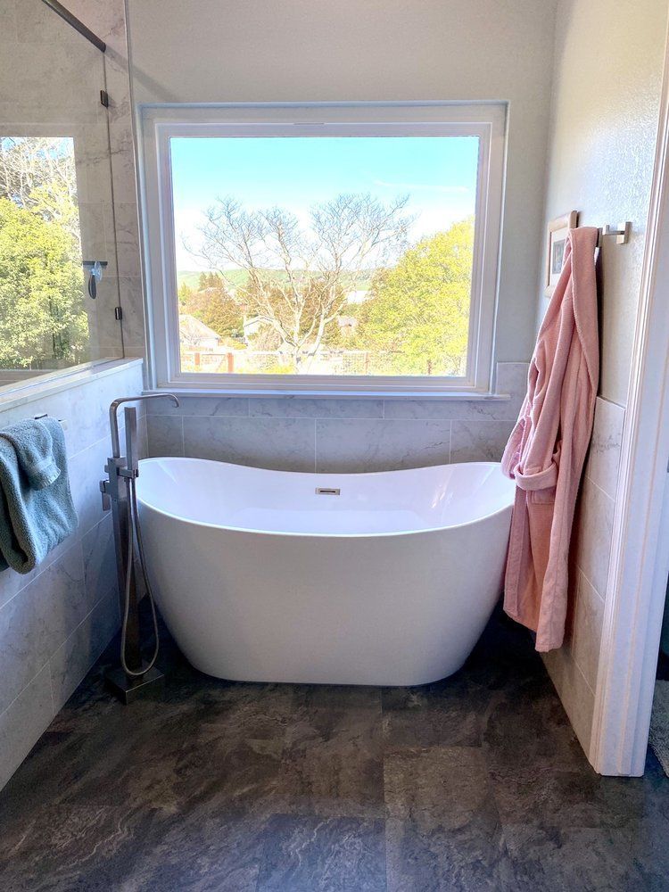 Bright bathroom with a freestanding white tub beneath a large window and pink towel on the right.