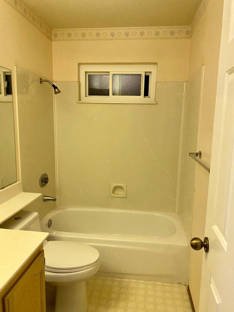 Small beige bathroom with bathtub, toilet, vanity, and window.