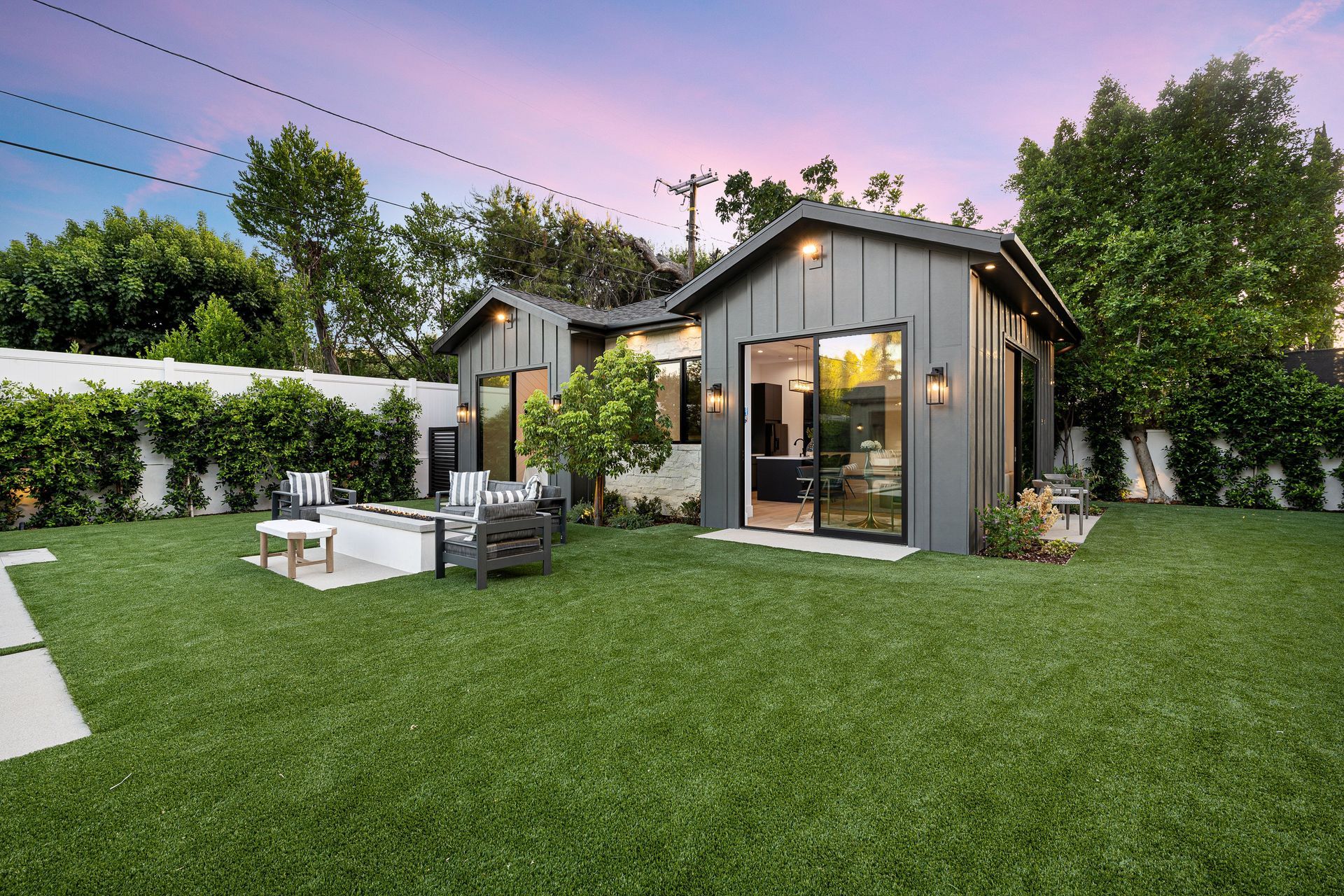Modern backyard with a small gray cottage, patio seating, and lush green lawn at dusk
