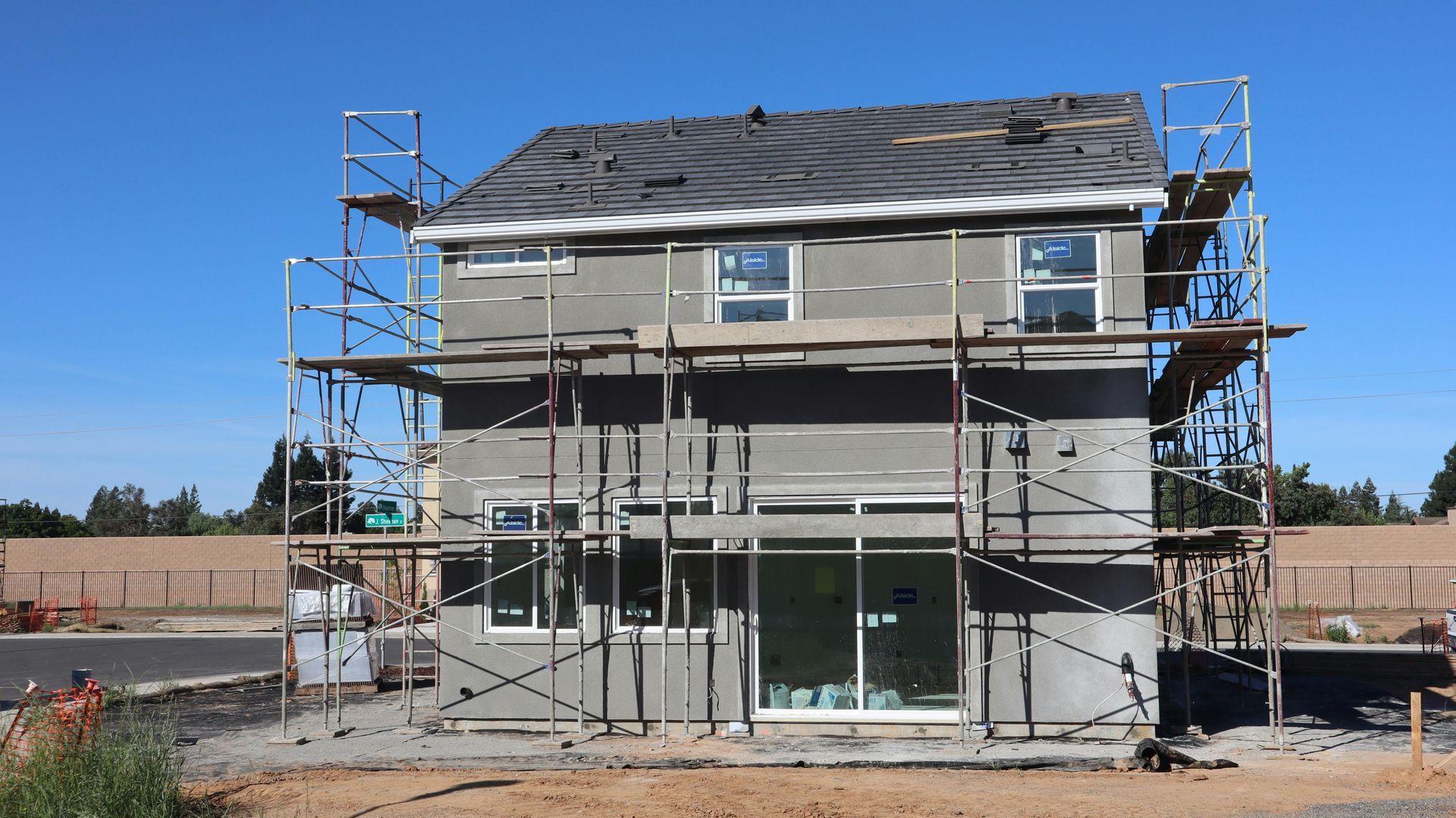 Two-story house under construction with scaffolding, gray siding, and unfinished roof under a clear blue sky