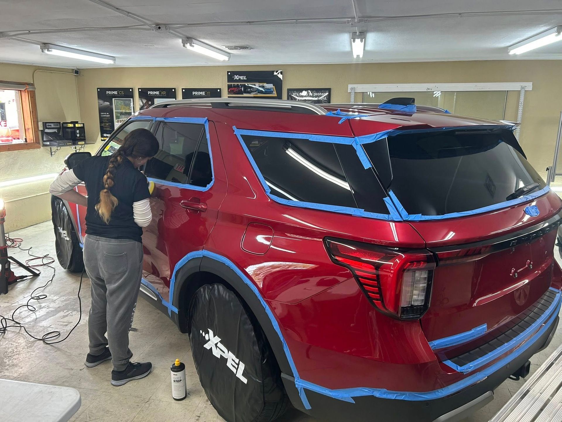 Woman working on a red SUV in a garage, vehicle masked with blue tape, wheels covered.