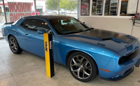 Blue Dodge Challenger with black hood, parked inside a window tinting shop.