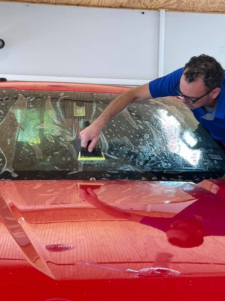 Man applies window tint to a red car windshield inside a garage using a squeegee.