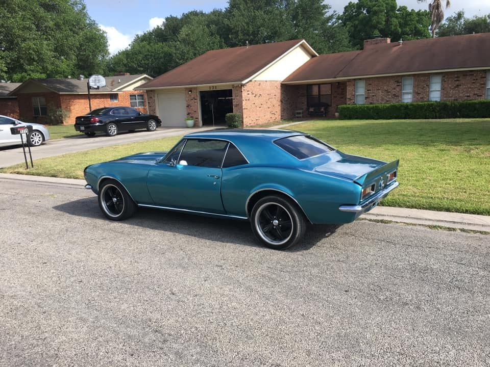 Blue classic Camaro parked on a residential street; houses and trees in background.