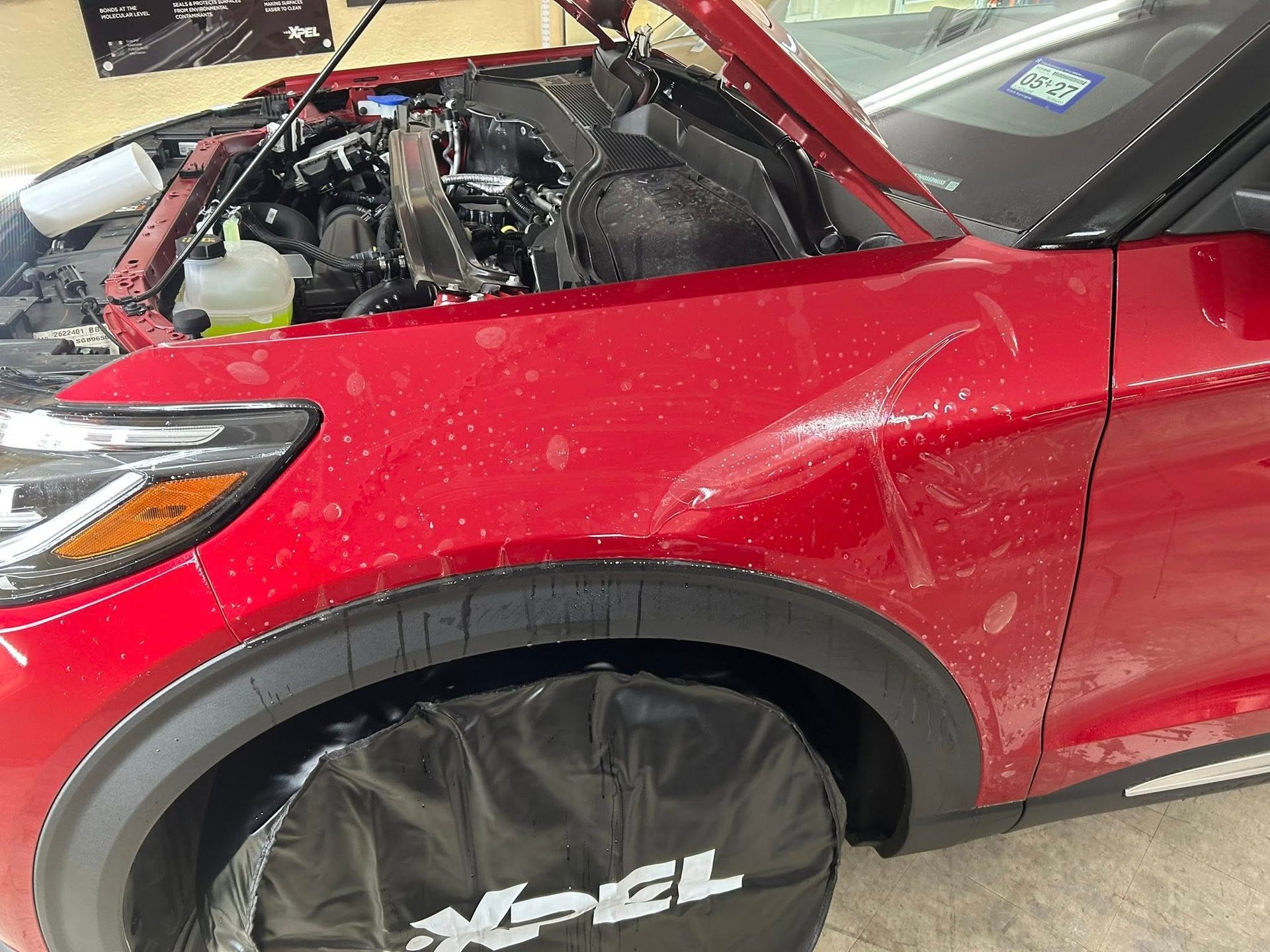 Red car with hood up, being worked on, covered in water drops. The Xpel logo is visible.