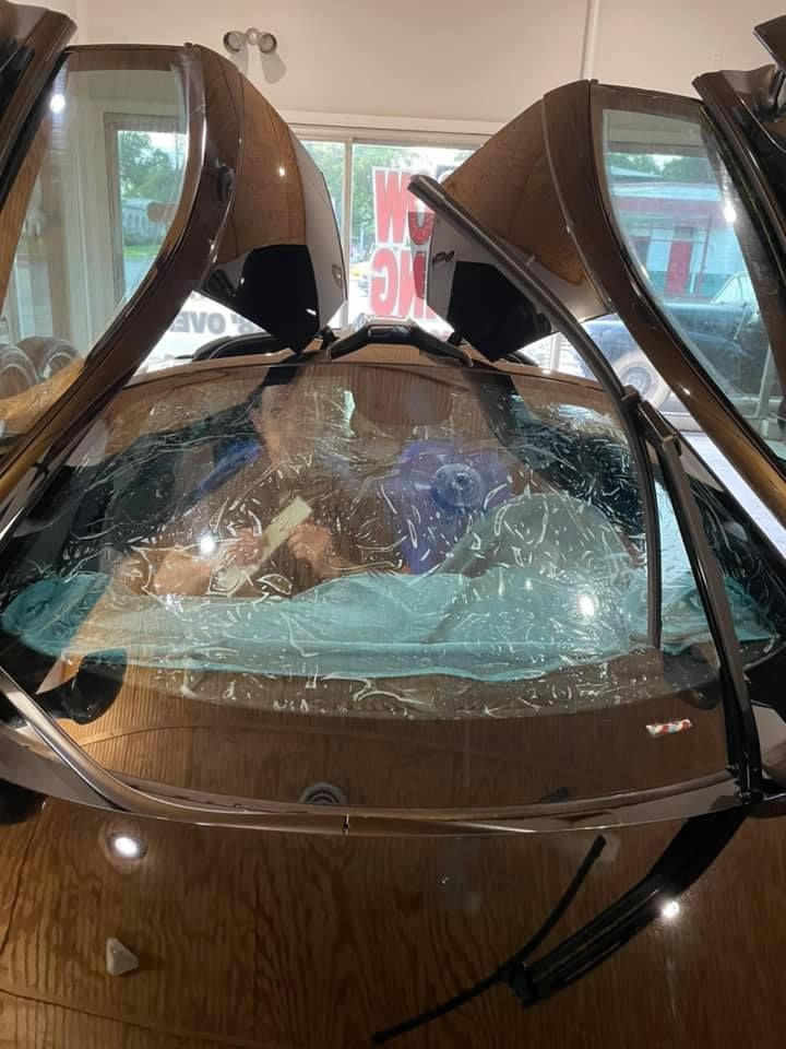 Person inside a clear, enclosed car-like structure, possibly a display, reading with opened doors.