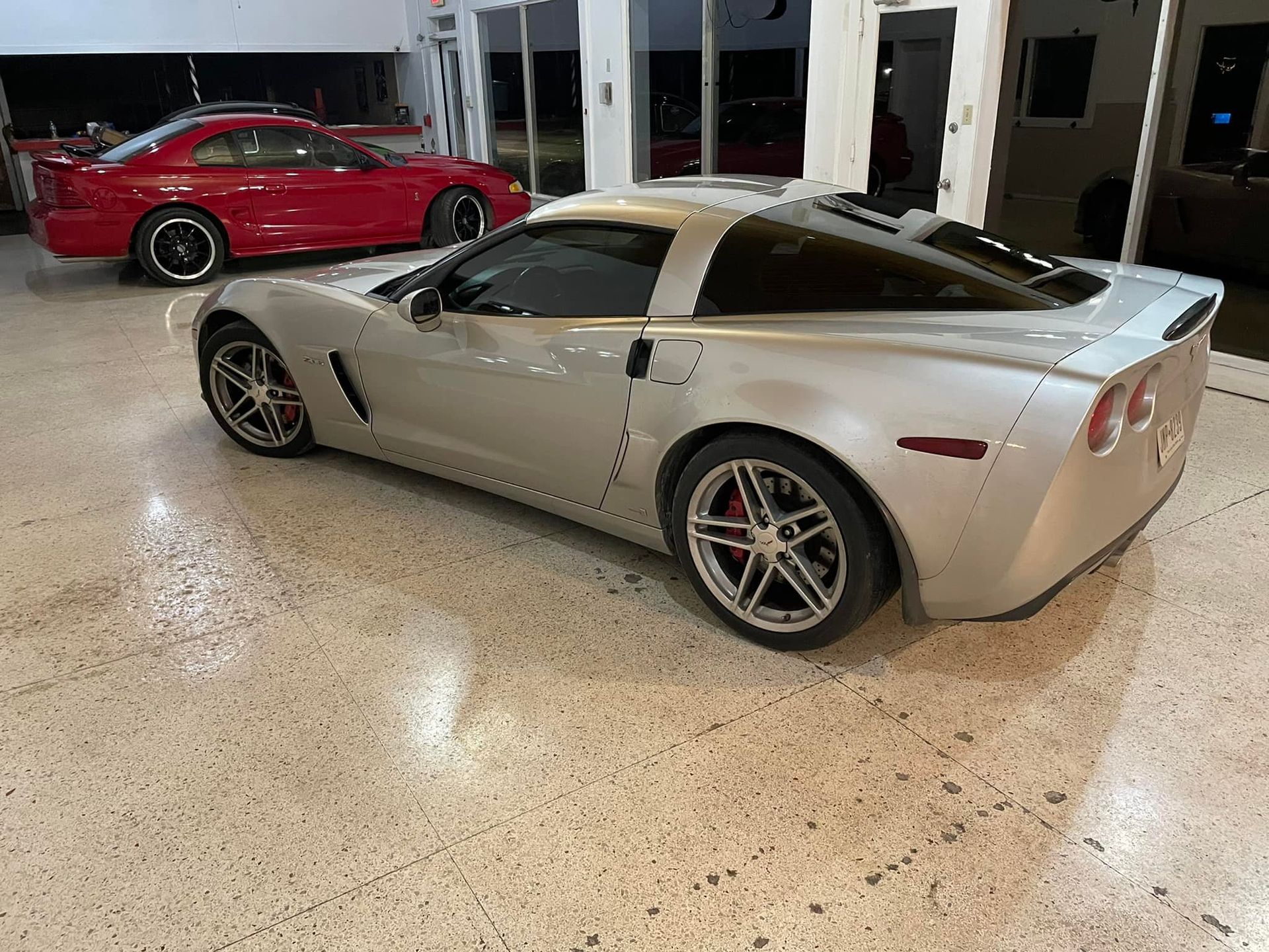 Silver Corvette parked indoors with a red Mustang behind it.