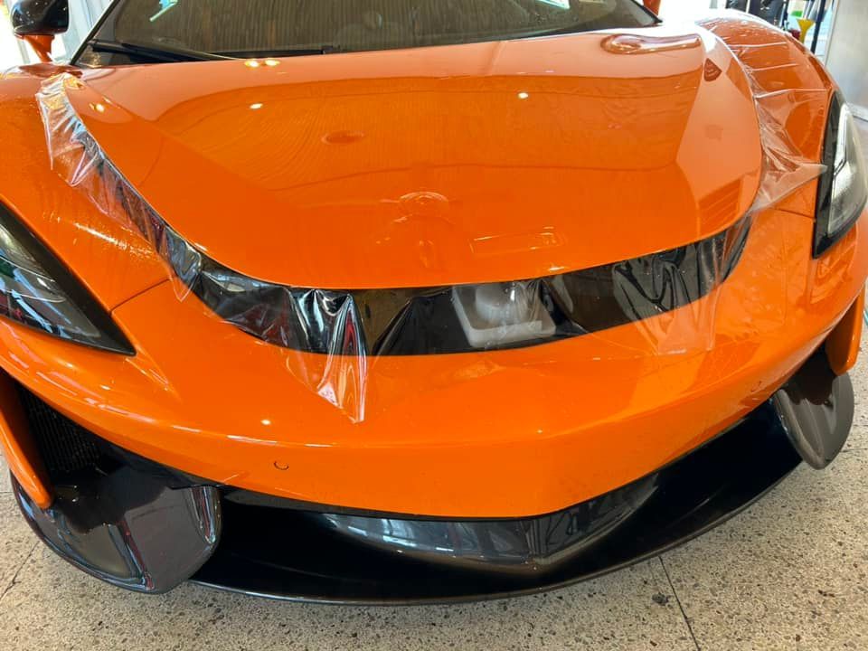 Orange sports car with protective film on the front. Black accents, indoor setting.