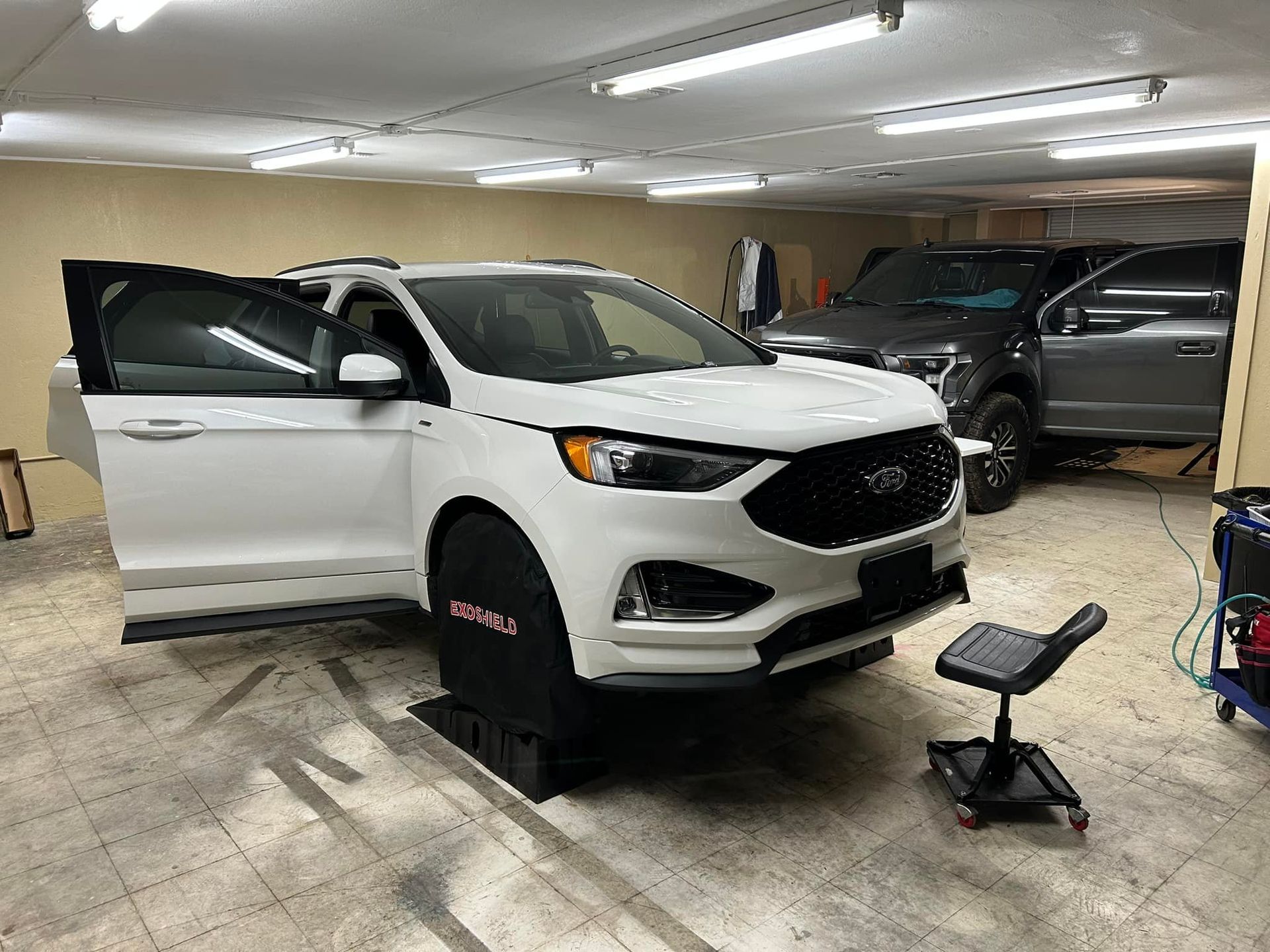 White Ford Edge with open door in a garage; a gray truck is in the background, and a rolling stool sits nearby.