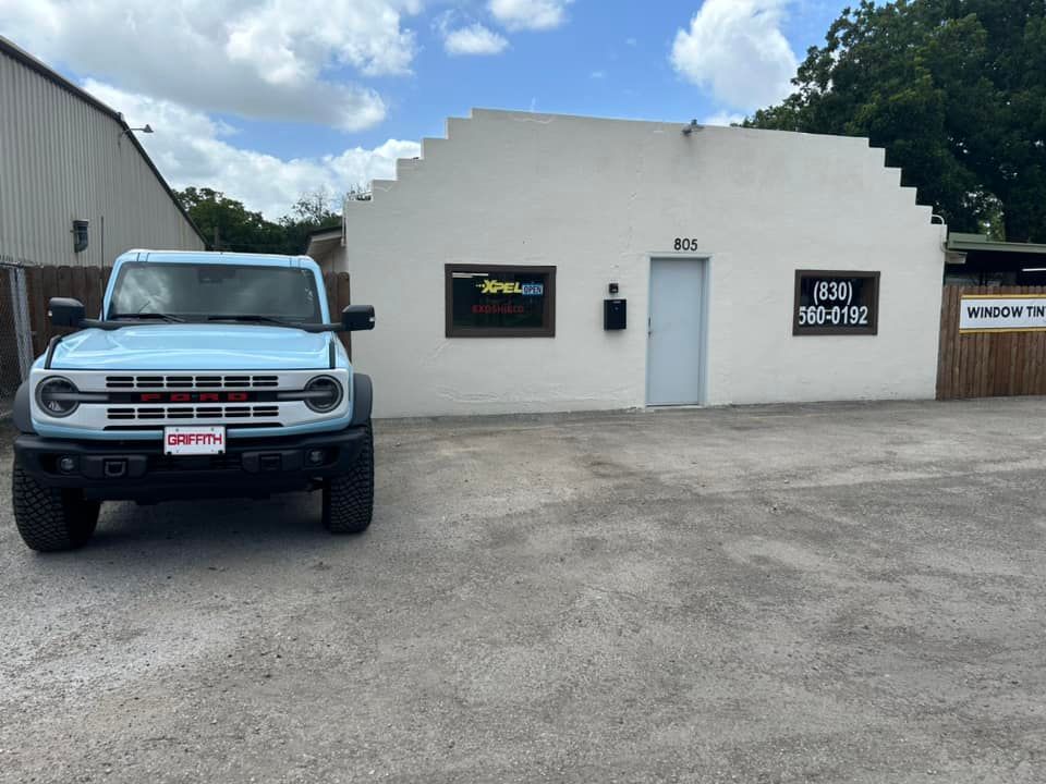 Light blue Ford Bronco parked in front of a white building with a business sign.