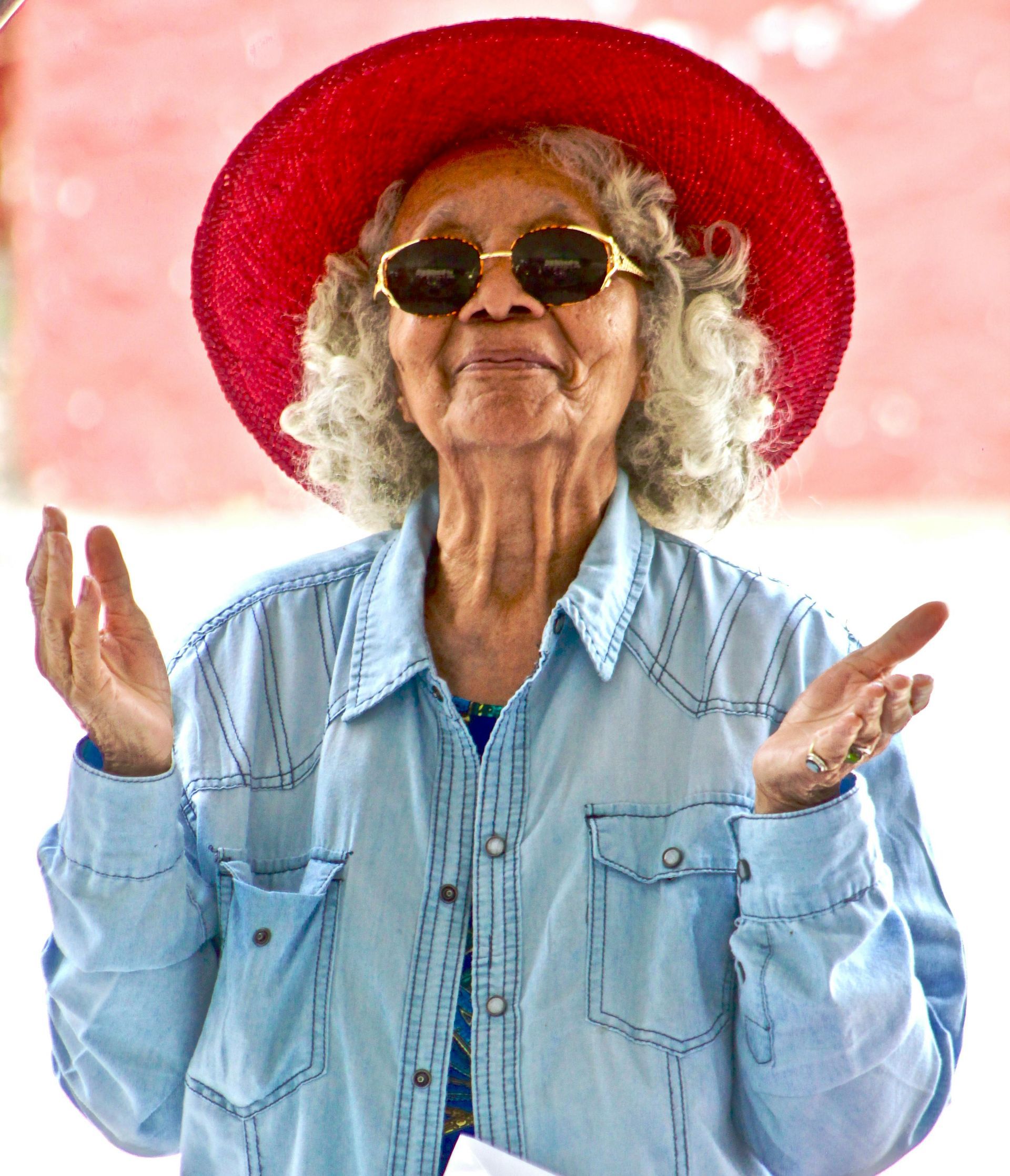 Older person in a red hat and sunglasses, smiling with raised hands in a light denim shirt.