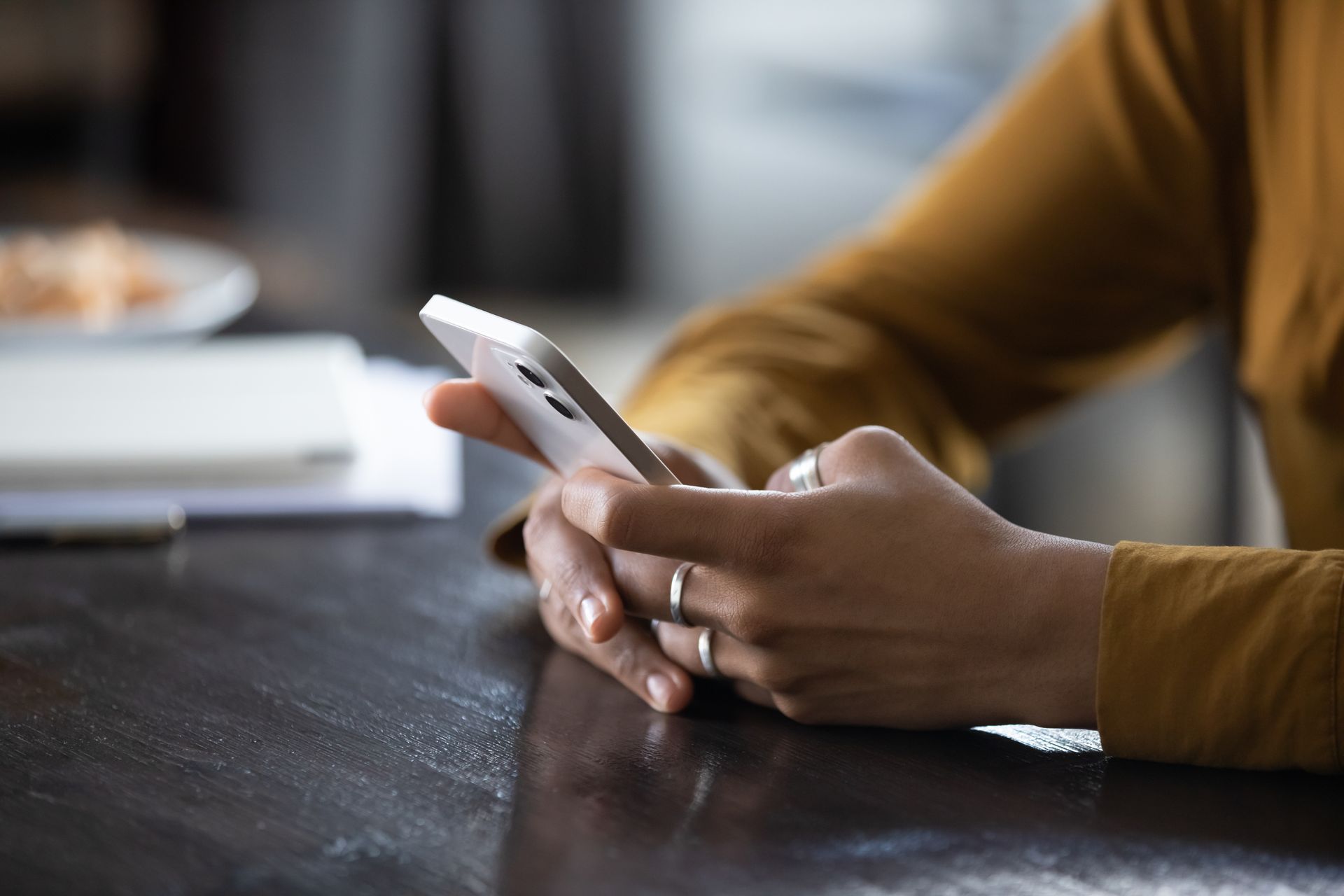 Hands holding a white smartphone at a dark wooden table in a softly blurred indoor setting