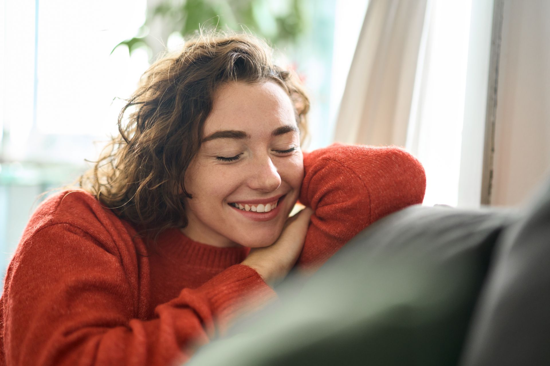 Smiling person in a red sweater resting by a window, with curly hair and eyes closed.