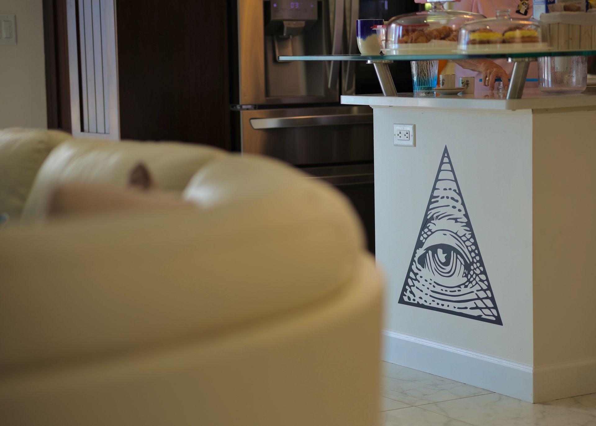 Cream-colored couch in foreground, kitchen island with eye-in-triangle symbol. Refrigerator and desserts in background.