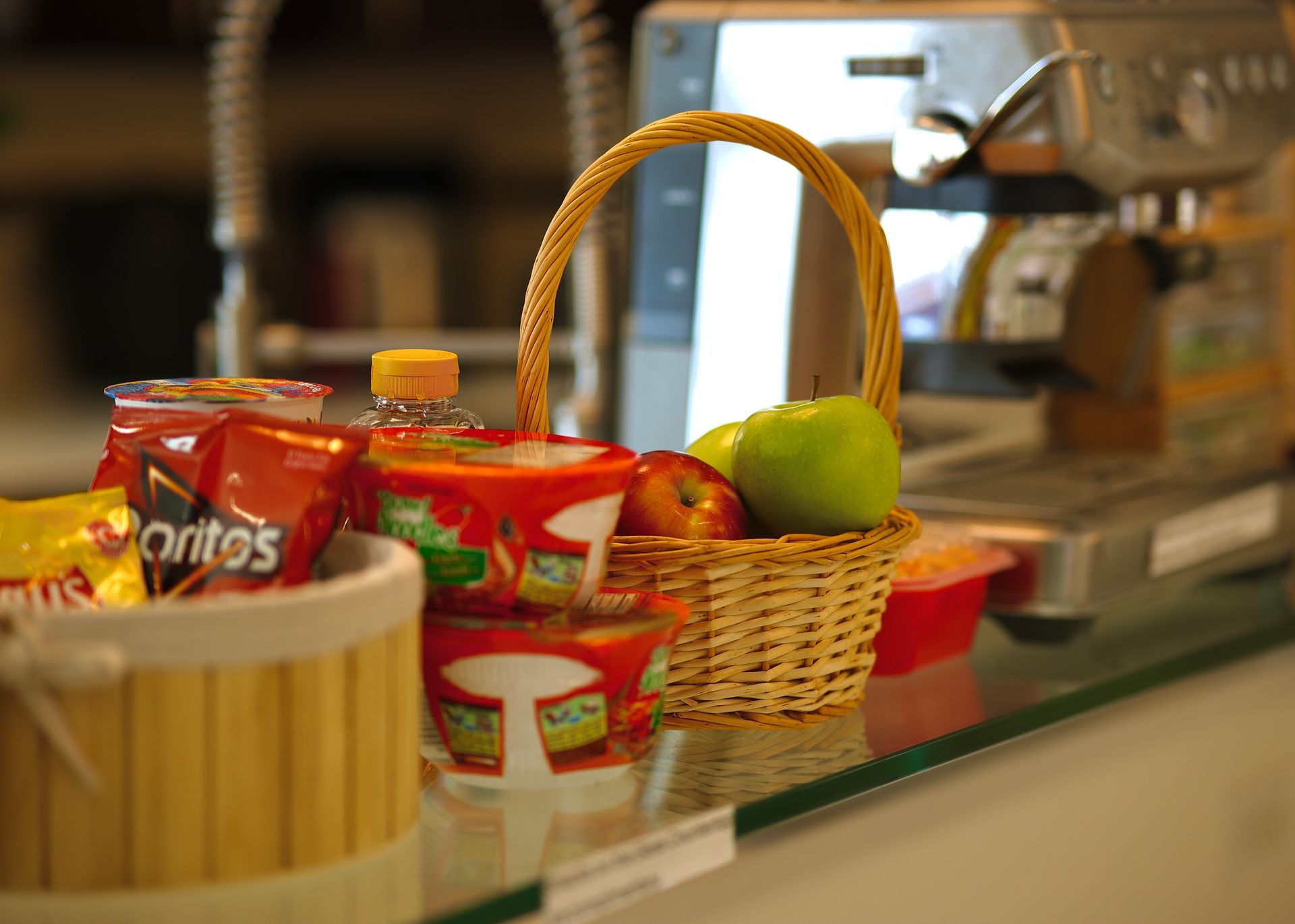 Basket of fruit next to snacks on a countertop; espresso machine in background.