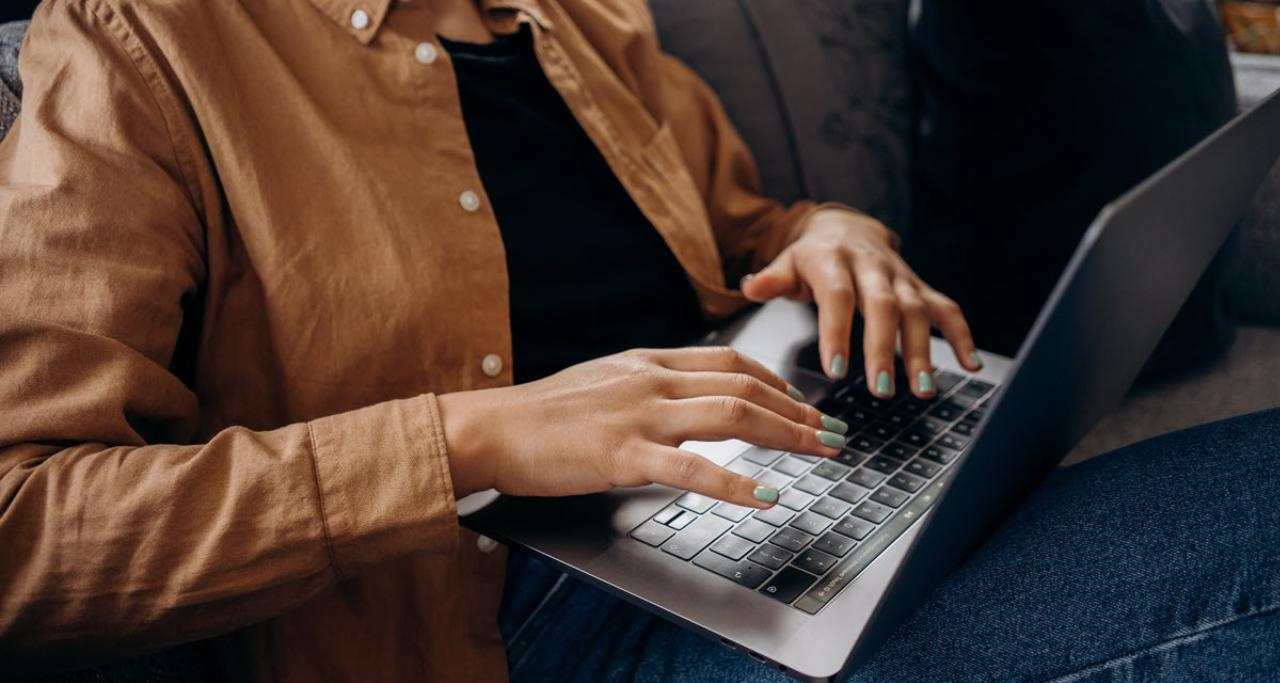 A person in a brown shirt typing on a laptop while sitting on a couch.