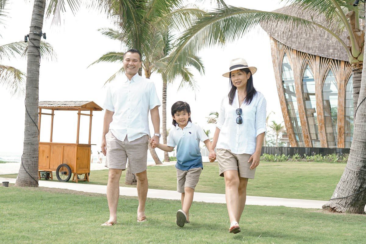 A family walks across a green lawn near a wooden food cart and tropical palm trees on a bright day.