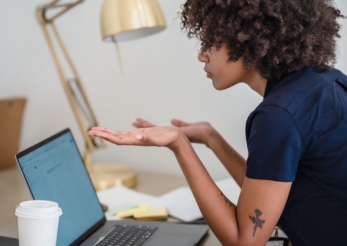 Person seated at a desk, gesturing toward a laptop in a home office.