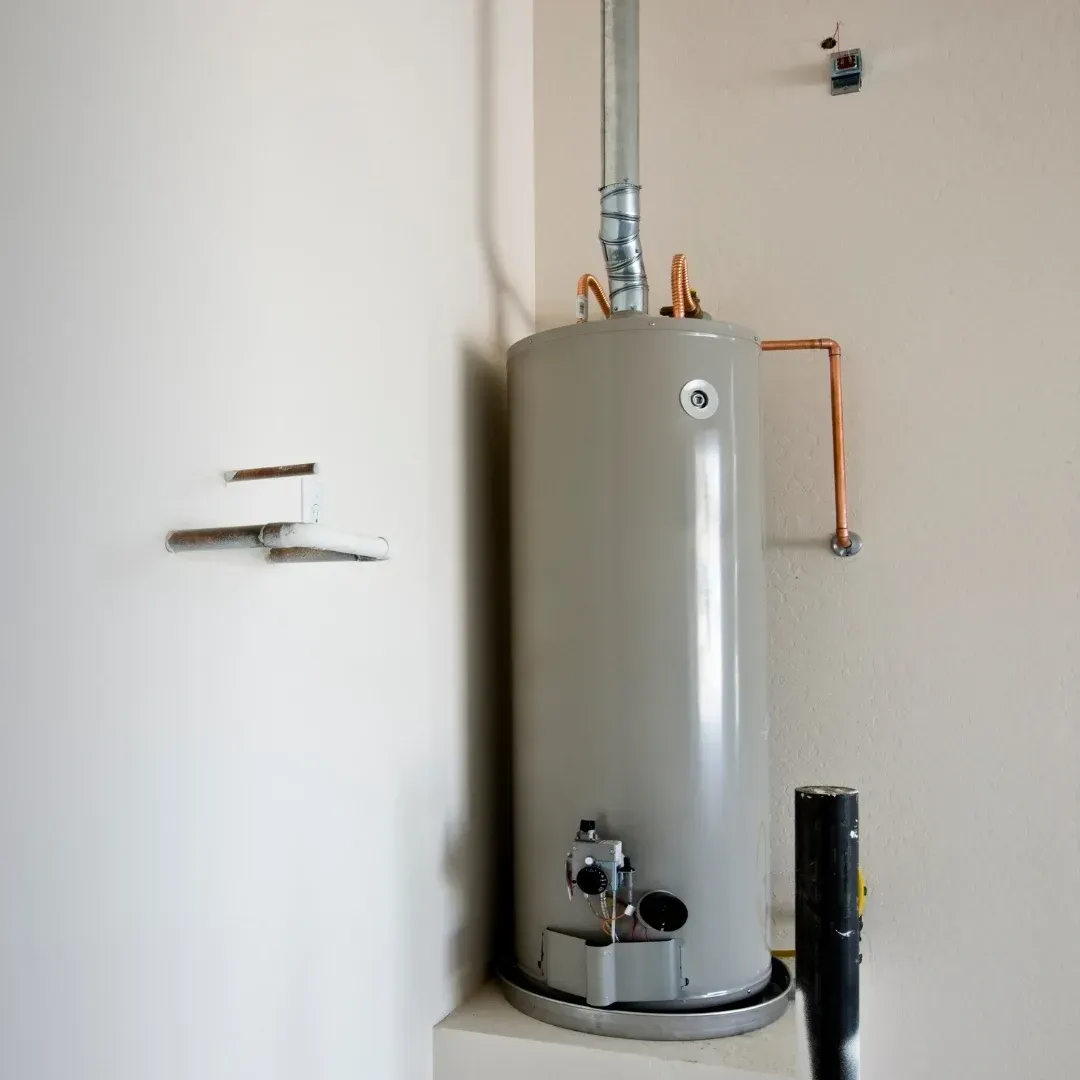 Gray water heater tank in a utility room with exposed pipes and a wall-mounted vent.