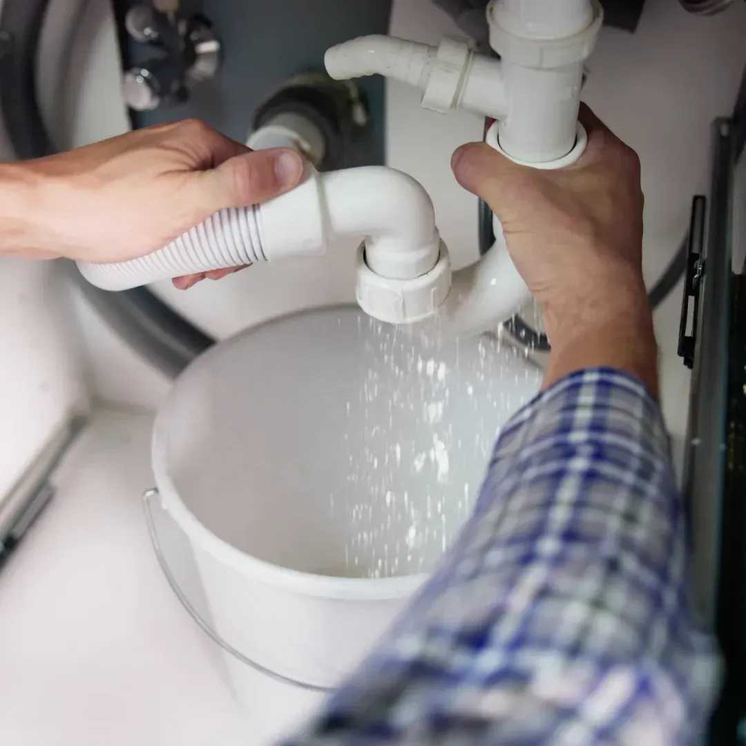 Hands fixing a white sink drain while water pours into a bucket underneath