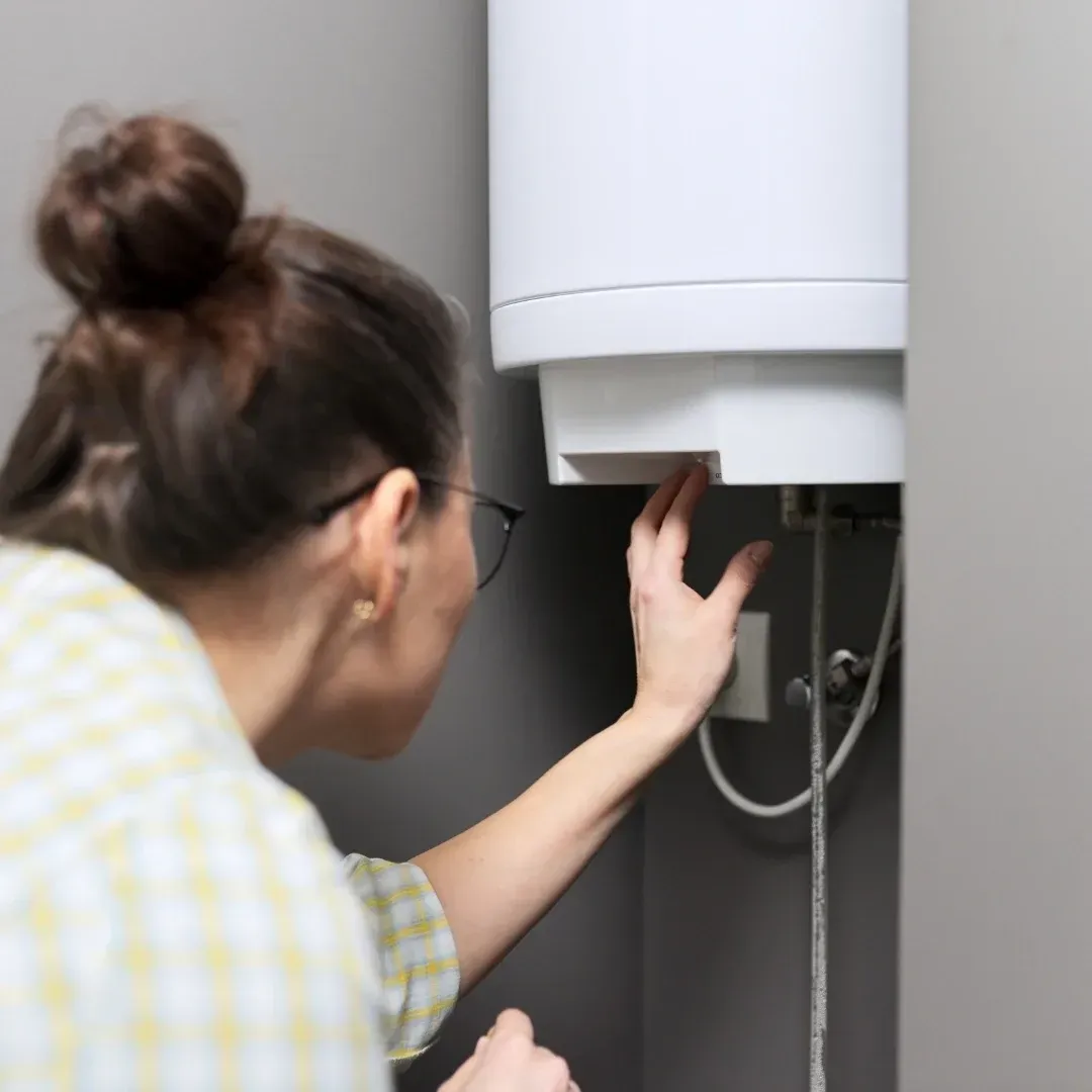 Person inspecting the underside of a white wall-mounted water heater with exposed pipes