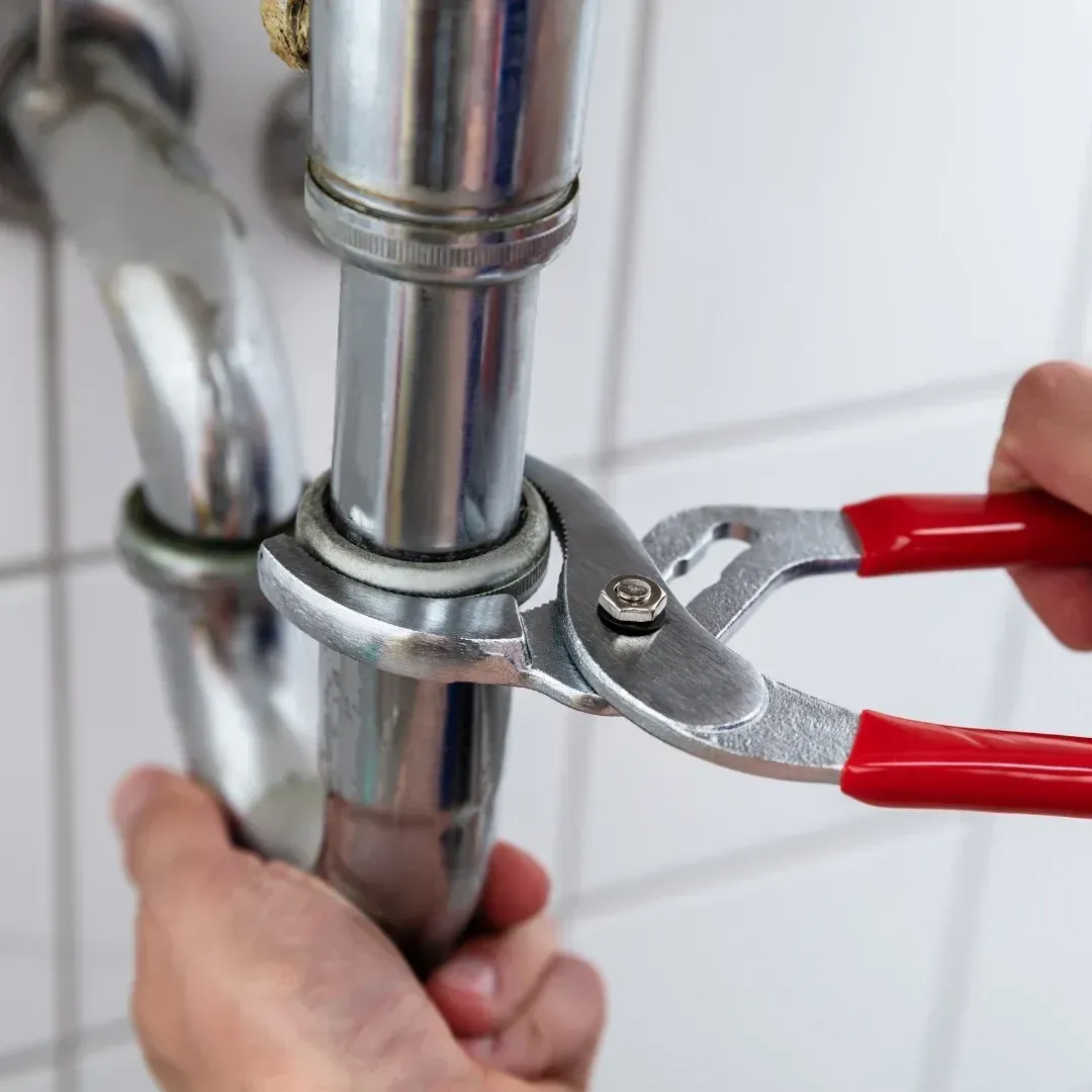 Hands using red-handled pliers to tighten a metal pipe clamp on a chrome plumbing pipe.