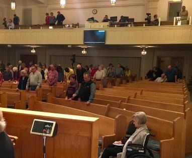 People standing in a church sanctuary with wooden pews and a balcony, viewed from the pulpit area