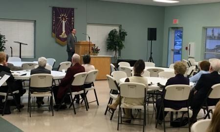 Speaker addressing seated audience in a community room with a podium and banner on the wall