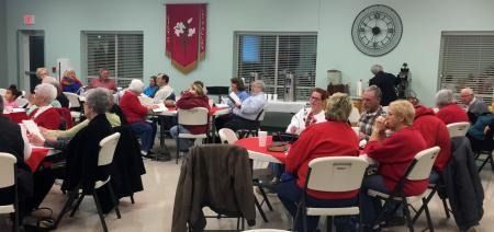 People seated at tables in a community hall, meeting or dining together under bright lights.