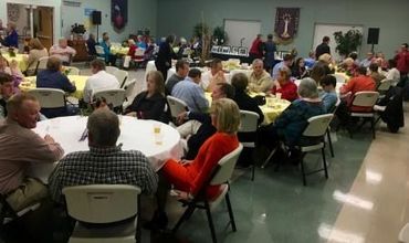Community gathering in a hall, with many people seated at round tables eating and talking.